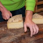 in primo piano mani maschili che tagliano una fetta da una mezza forma di formaggio.
