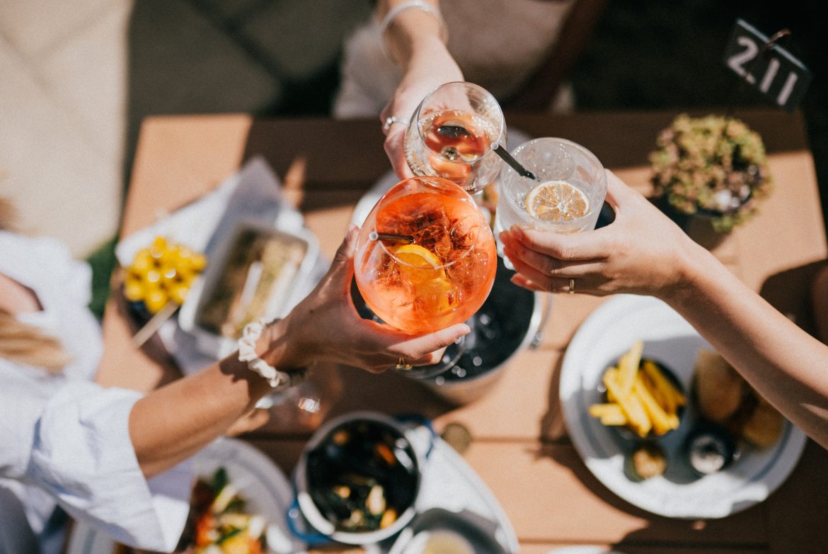Vista dall’alto di un aperitivo all’aperto: quattro persone brindano con cocktail e calici intorno a un tavolo con piatti e stuzzichini