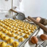 tortellini freschi disposti in file ordinate in una teglia. Sullo sfondo utensili da cucina e uova