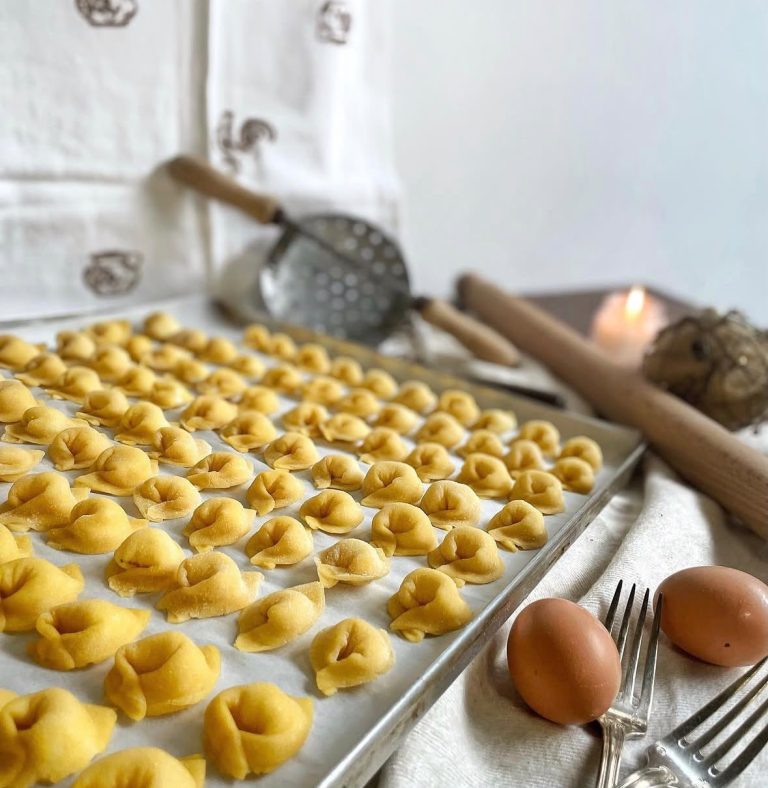 tortellini freschi disposti in file ordinate in una teglia. Sullo sfondo utensili da cucina e uova