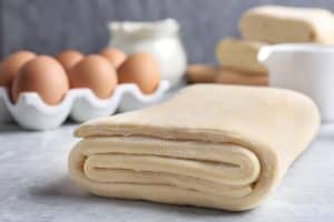 Impasto di pasta sfoglia piegato su un piano di lavoro, con uova e ingredienti caseari sullo sfondo.