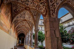 portico con arcate riccamente affrescate sui toni dell'ocra e del crema. Sullo sfondo un cielo azzurro e terso. 