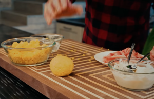 Una persona prepara il cibo con pane di mais, prosciutto e panna acida su un tagliere di legno.