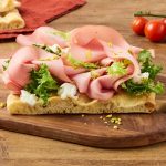 In primo piano una pinsa riccamente farcita con mortadella, burrata, insalata verde e granella di pistacchi, servita su un tagliere in legno.