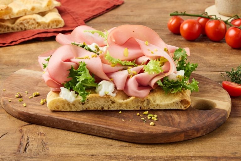 In primo piano una pinsa riccamente farcita con mortadella, burrata, insalata verde e granella di pistacchi, servita su un tagliere in legno.